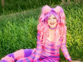 The Cheshire Cat lounging in the Calgary grass with the forest behind her, looking out for the Red Queen of Hearts